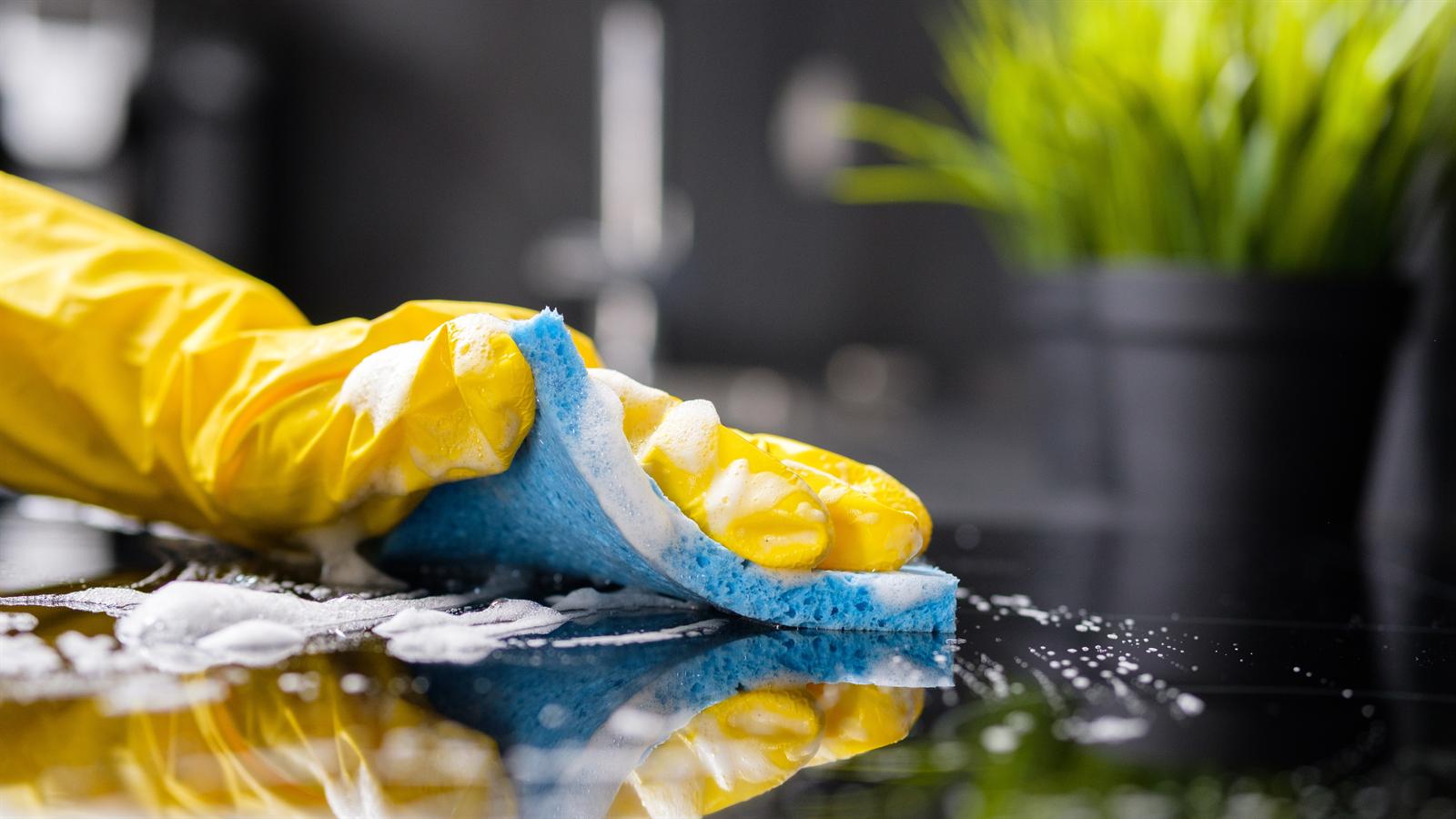 A gloved hand scrubbing a dark kitchen countertop with a soapy sponge