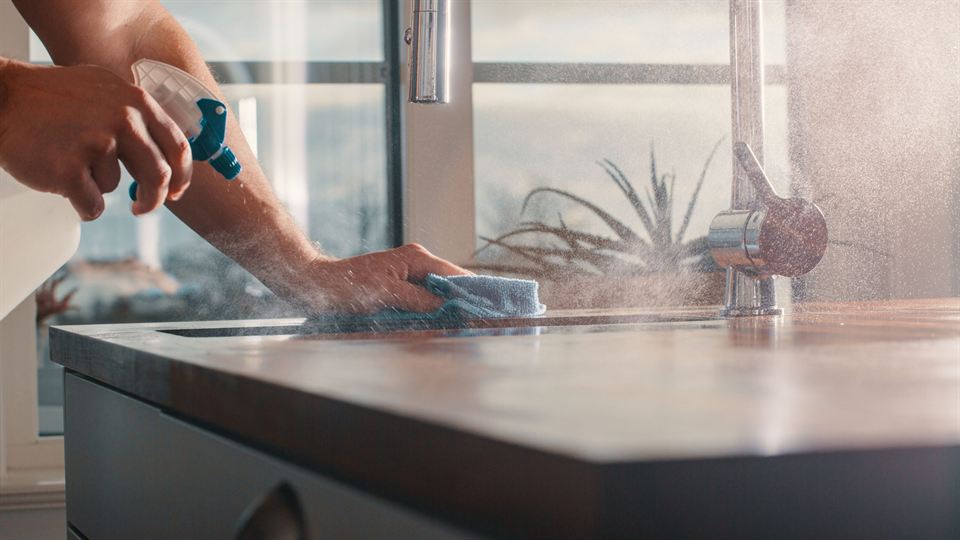 A cleaner spraying and wiping a kitchen countertop by a bright window
